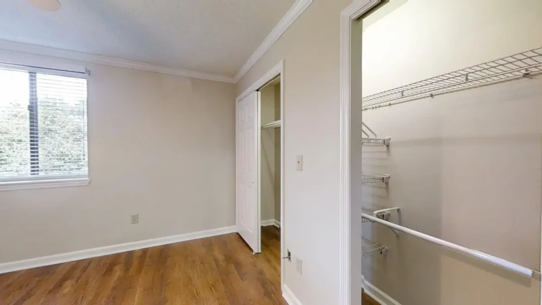 Empty bedroom with hardwood floor, window with blinds, and an open closet with wire shelves.