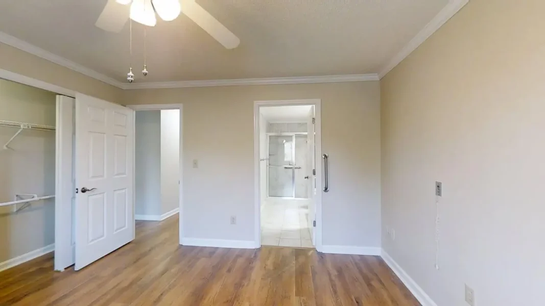 Empty bedroom with light wood floor, open closet on the left, and a doorway leading to a bathroom with a glass shower.