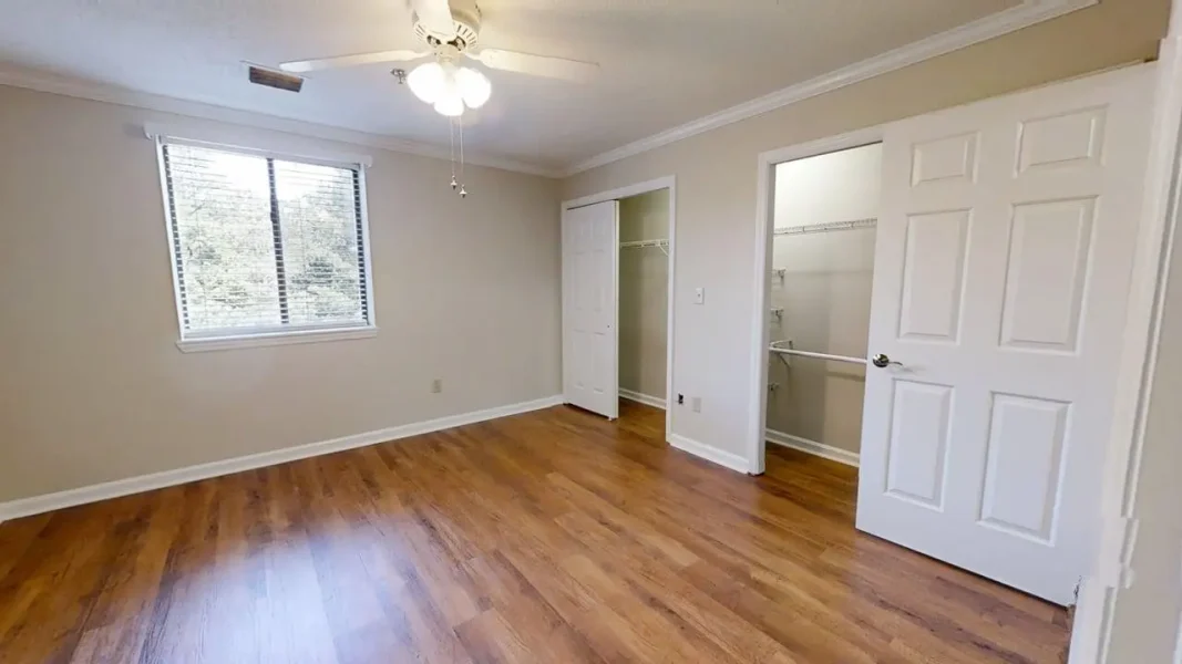 Empty bedroom with light hardwood flooring, beige walls, a ceiling fan, and a large window letting in natural light.