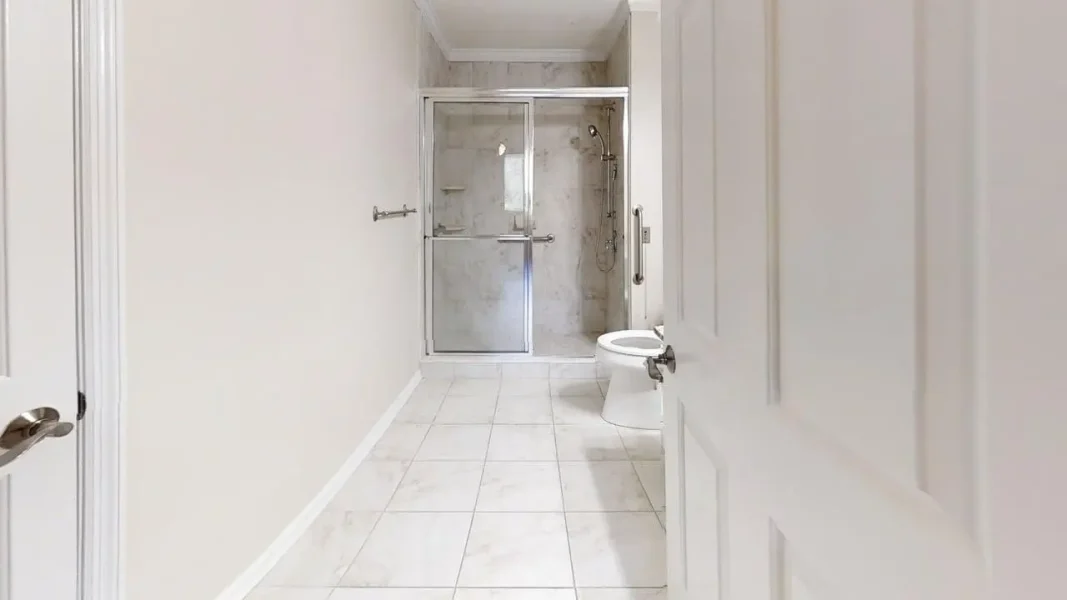Narrow white-tiled bathroom with a glass-enclosed shower at the far end and a toilet on the right.