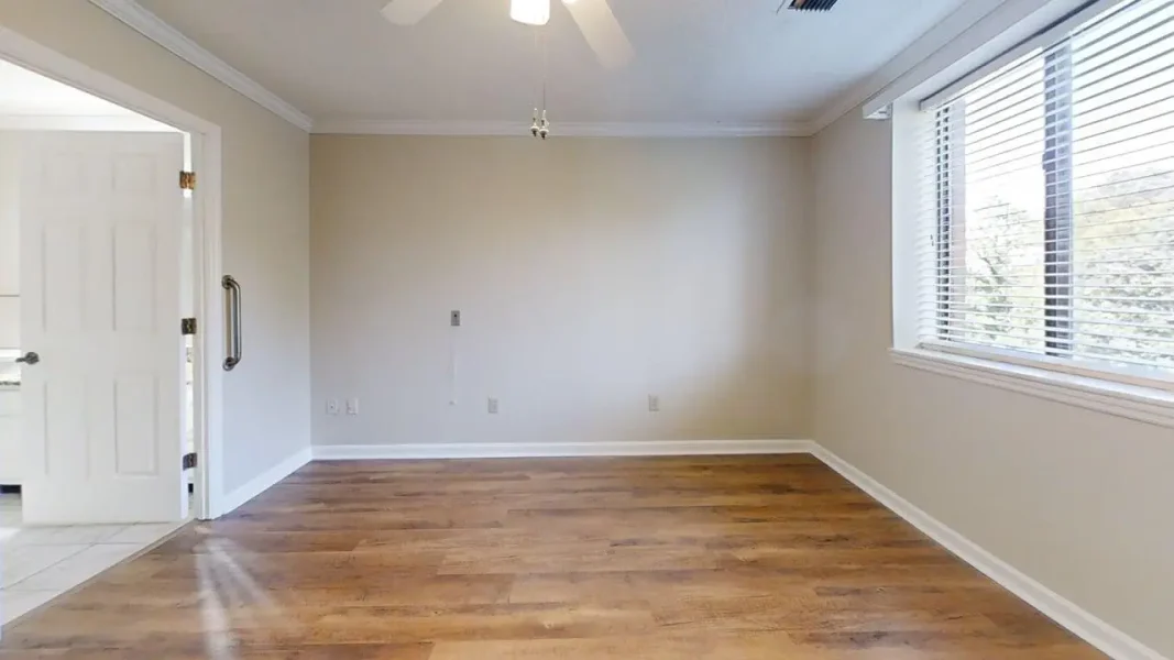 Empty living room with light hardwood floors, beige walls, and a large right-side window with blinds letting in natural light, plus an open door leading to another room on the left and a ceiling fan overhead.