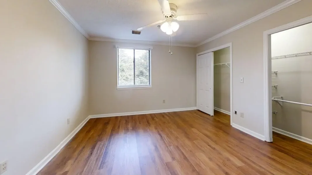 Empty bedroom with hardwood floors, beige walls, white trim, and a ceiling fan. A window provides natural light; a closet with doors is visible on the right.