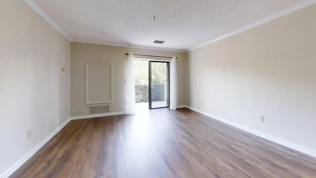 Empty living room with light beige walls, white baseboards, and wood-look flooring; sliding glass door with sheer white curtains opens to a balcony.