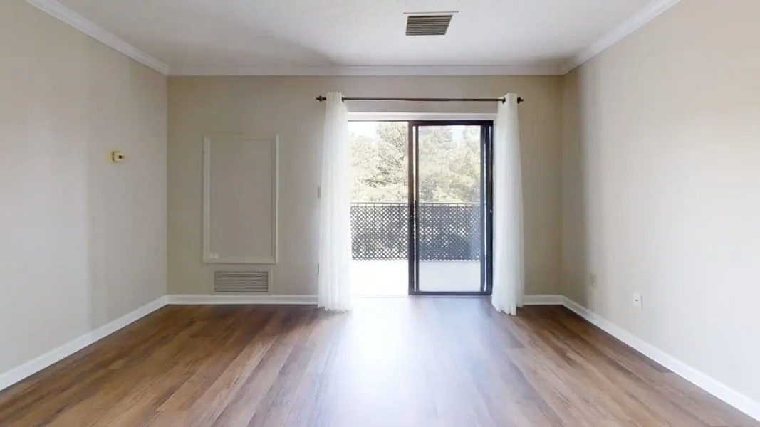 Empty living room with beige walls, white baseboards, and dark wood flooring, featuring a sliding glass door with sheer white curtains to a balcony.