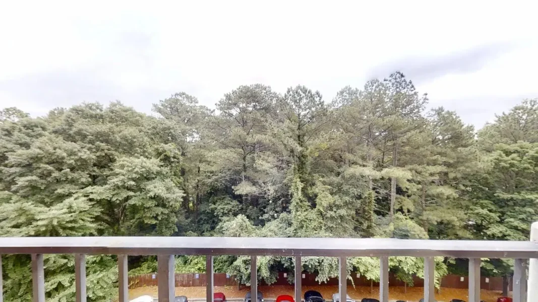 Balcony railing with a view of dense green trees and a parking area below under a cloudy sky.