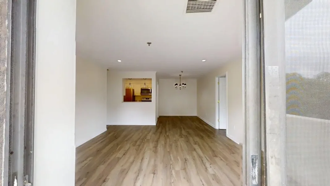 Empty open-plan living space with wood-like flooring, a kitchen pass-through, and a dining chandelier in the back corner.