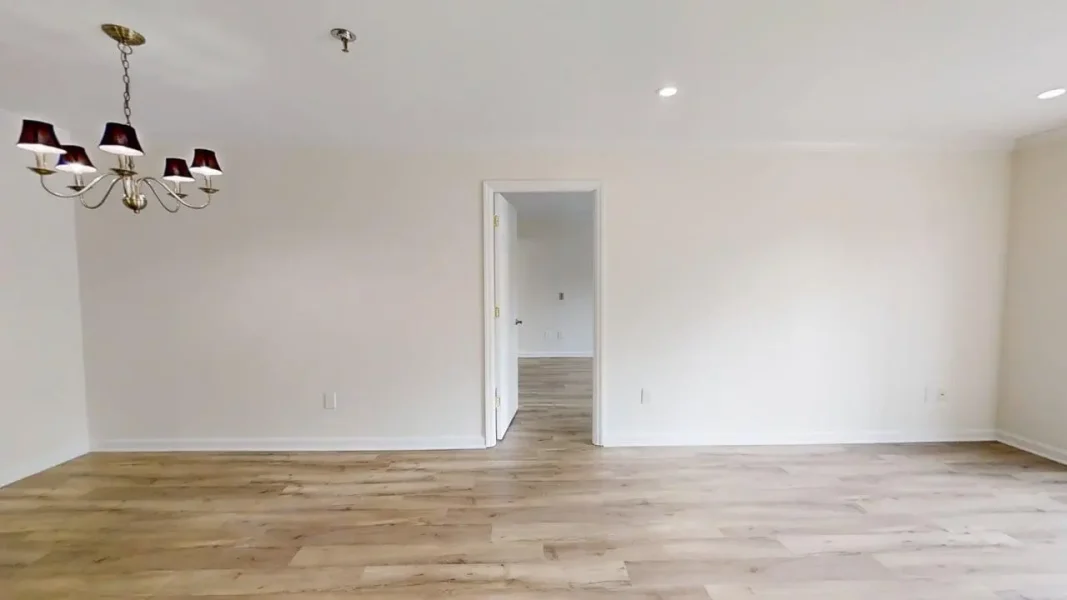 Empty living area with light wood floors, white walls, and a chandelier hanging on the left; doorway in the center leads to another room.