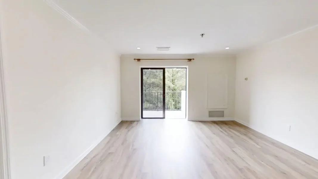 Bright empty living room with light wood flooring, white walls, and a sliding glass door to a balcony showing greenery outside.