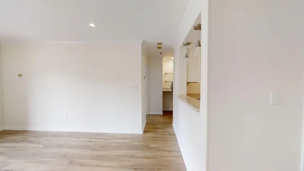Empty living area with light wood floors, white walls, and a kitchen pass-through on the right; hallway at the back center.