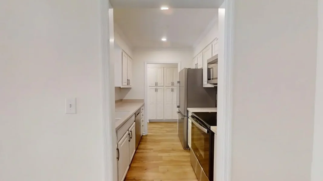 Bright galley kitchen with white cabinets, stainless steel fridge, range, and microwave; light wood floor.