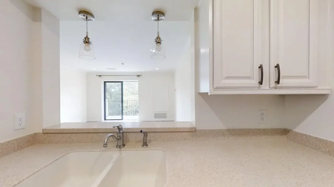 Kitchen counter with a sink and chrome faucet, white cabinetry, and pendant lights; view into a bright living area through a pass-through window.