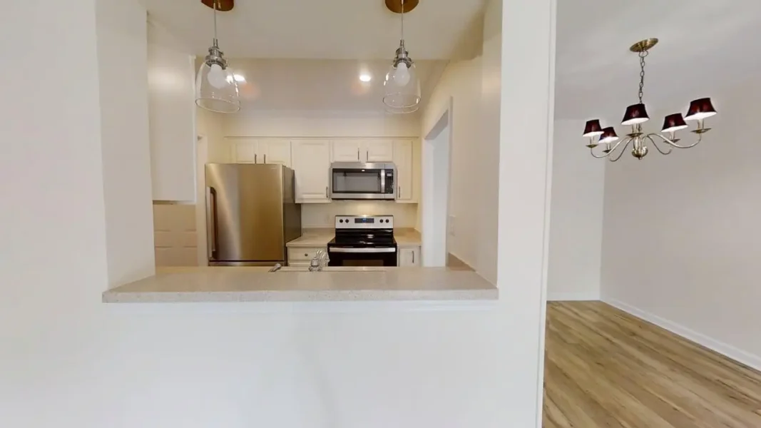 Open‑concept kitchen with white cabinets and stainless steel fridge, stove, and microwave; pass‑through counter to a dining/living area with a chandelier and wood floor nearby.