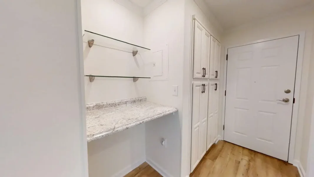 White mudroom with granite countertop, glass wall shelves, and tall white cabinets along a wall near a white entry door; wood-tone floor.