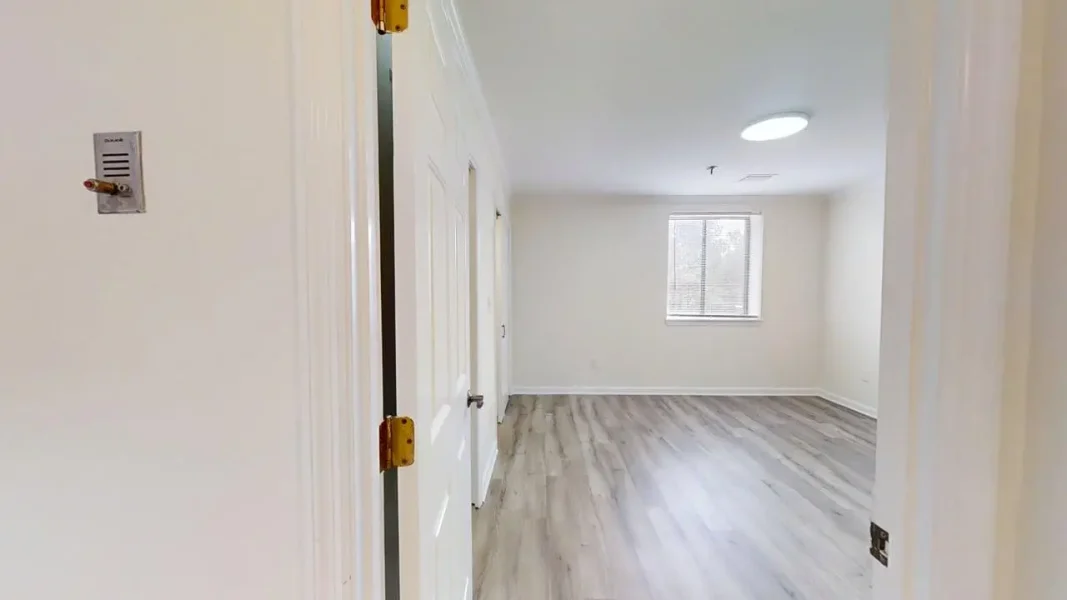 Bright, empty living area with light gray wood-look flooring, white walls, a window with blinds, and a round ceiling light fixture.