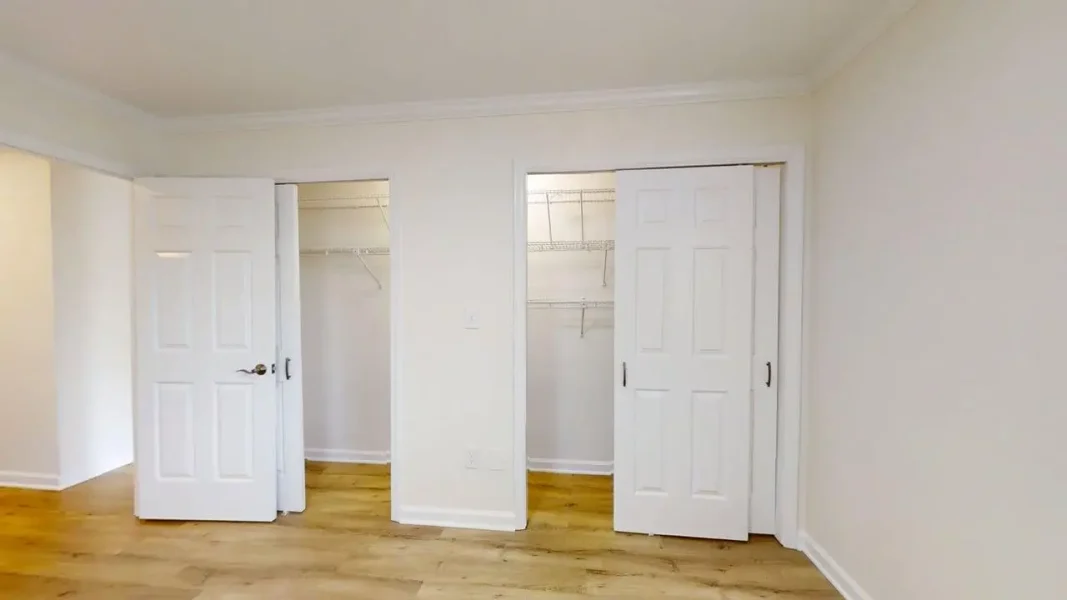 Empty white room with light wood floor; left closet door open showing wire shelves, right closet doors closed.