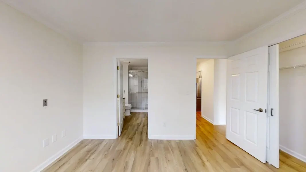 Empty bedroom with light wood floors and white walls; doorway to a bathroom on the left and an open closet on the right.