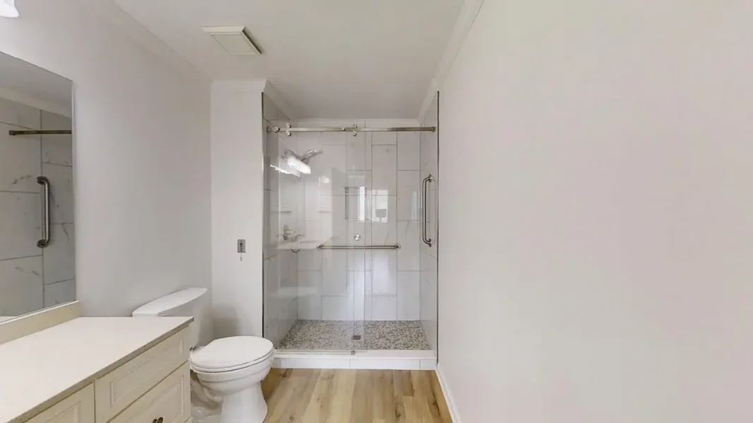 Modern bathroom with a glass-enclosed shower, white tiles, toilet, and beige vanity on light wood flooring in a bright space.