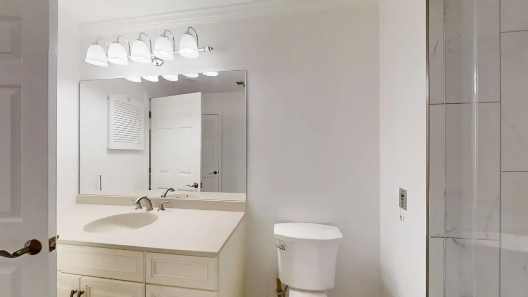 White bathroom with a single vanity, large mirror, and a toilet beside a glass shower.