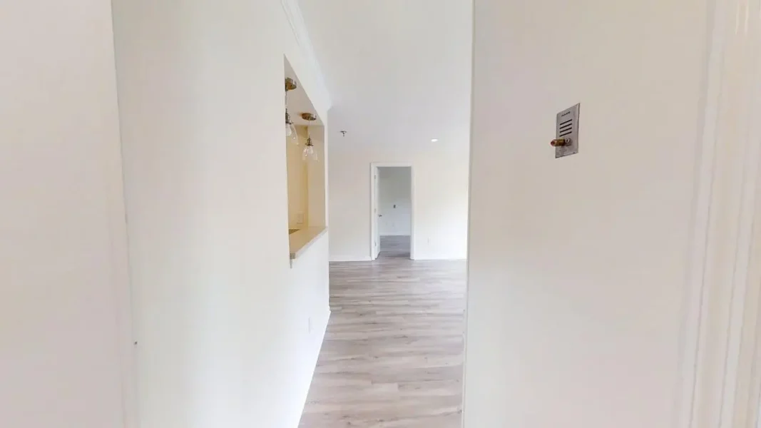 Bright, white hallway with light gray wood floors, a kitchen pass-through on the left, and a doorway at the far end.