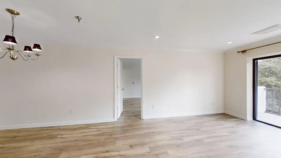Bright empty living room with light wood floors, white walls, and a chandelier on the left; sliding glass door to a balcony on the right; doorway to another room in the center.
