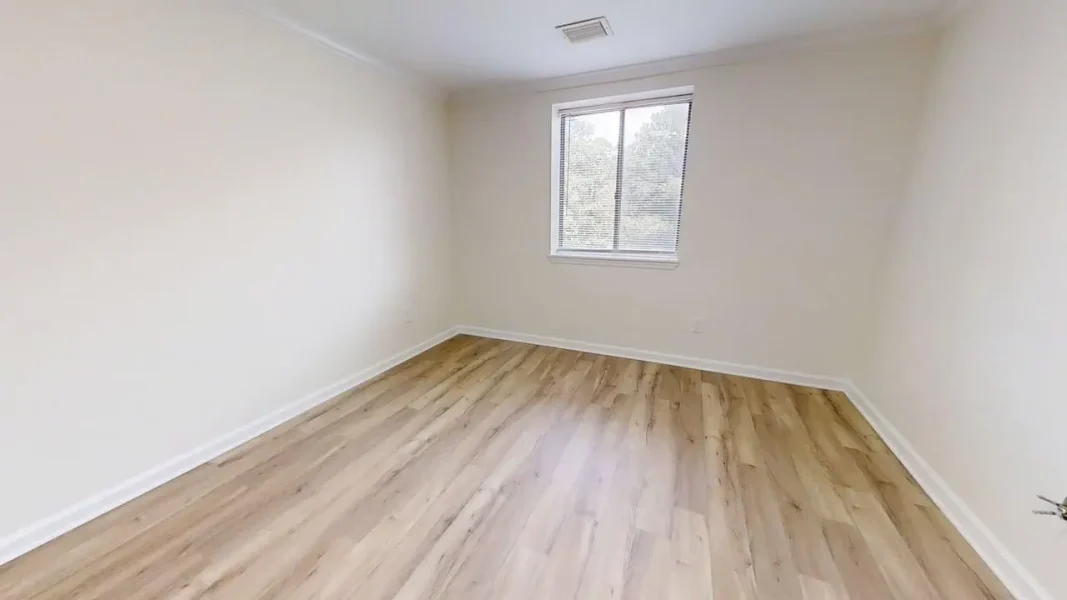 Empty rectangular room with light wood laminate flooring, white walls, baseboard trim, and a single window letting in natural light.