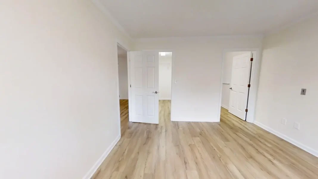 Empty room with light wood flooring and white walls, two open doors leading to adjacent spaces.