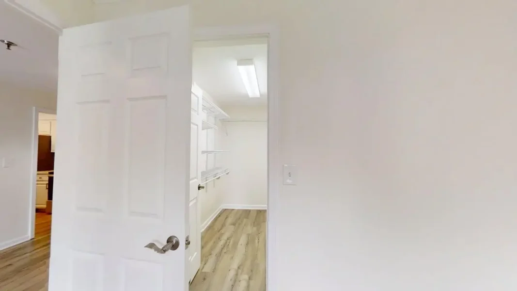 Open doorway to a bright walk-in closet with white built-in shelves and hanging rods, wood-look flooring visible in a minimalist home interior.