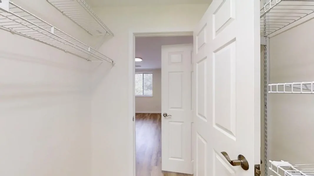 Empty walk-in closet with white wire shelves on both sides and an open door leading to a room with wood flooring.
