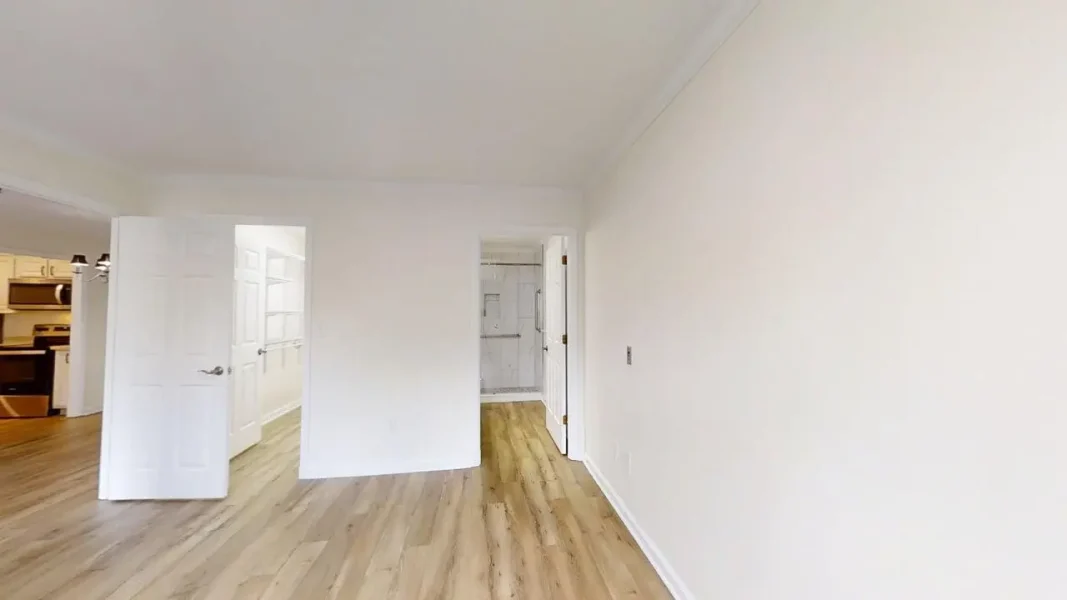 Vacant living space with light wood floors, white walls, and two doorways: to the left a kitchen and straight ahead a bathroom entry.