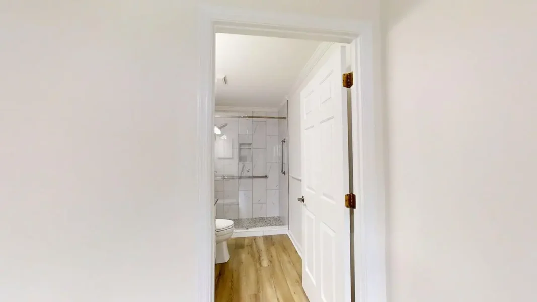 Open white door reveals a small, modern bathroom with wood-like floor, tiled shower enclosure, and grab bars near the wall behind a toilet.