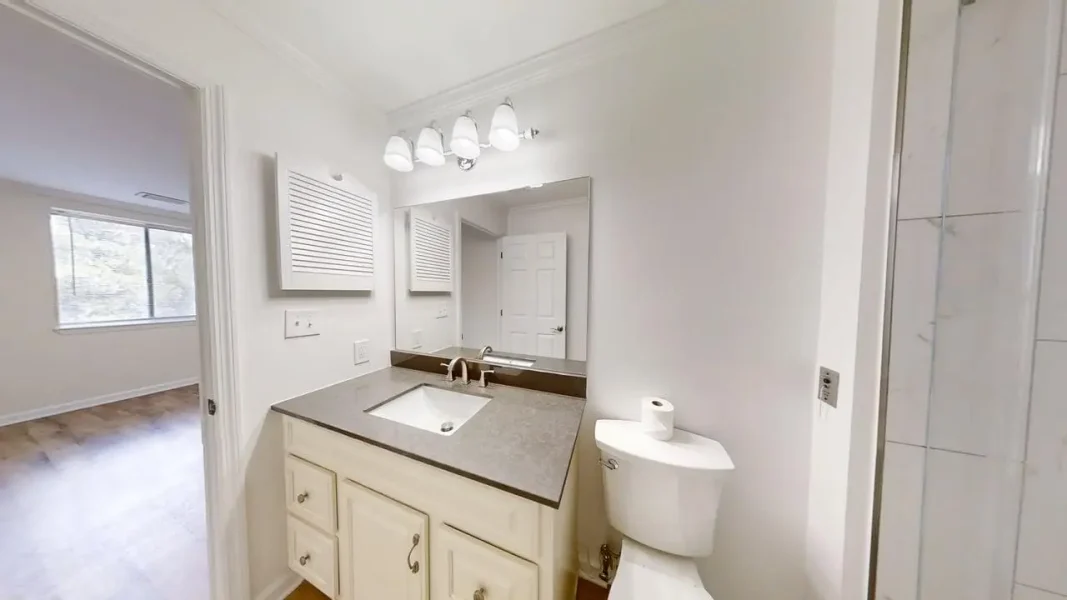 Bathroom vanity with gray countertop, white cabinets, large mirror, and toilet; shower visible on the right. Navigates to an adjoining room through an open door.