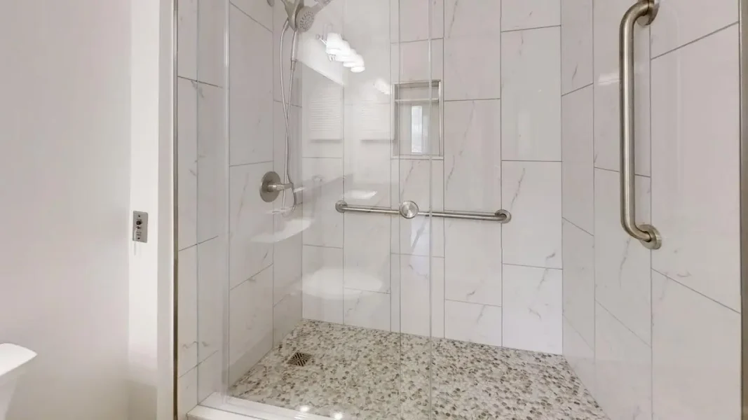 ADA-compliant shower with glass door, two grab bars, and handheld showerhead on a tiled wall. Underfoot is a textured, non-slip floor with a drain in the corner.