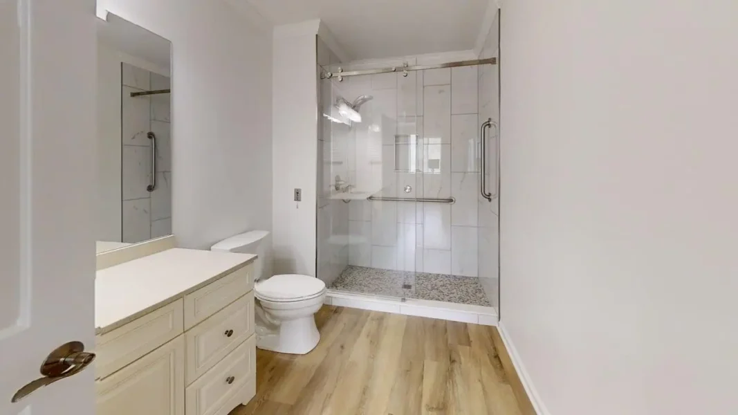 Bathroom with white vanity, mirror, toilet, and glass-walled shower featuring chrome grab bars and pebble-floor shower.