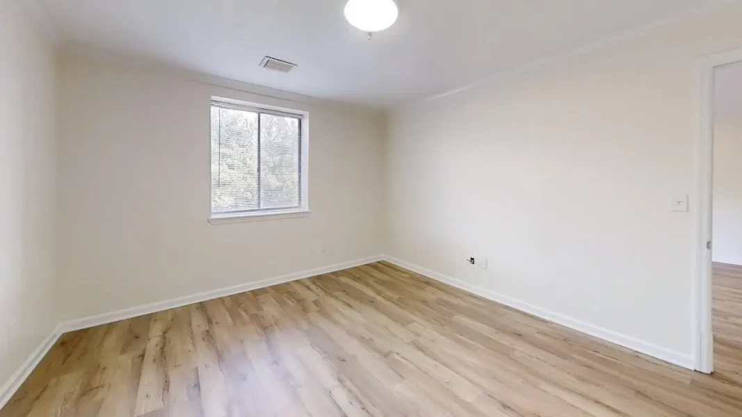 Empty beige-walled room with light wood flooring, a single window with blinds, and a ceiling light fixture.