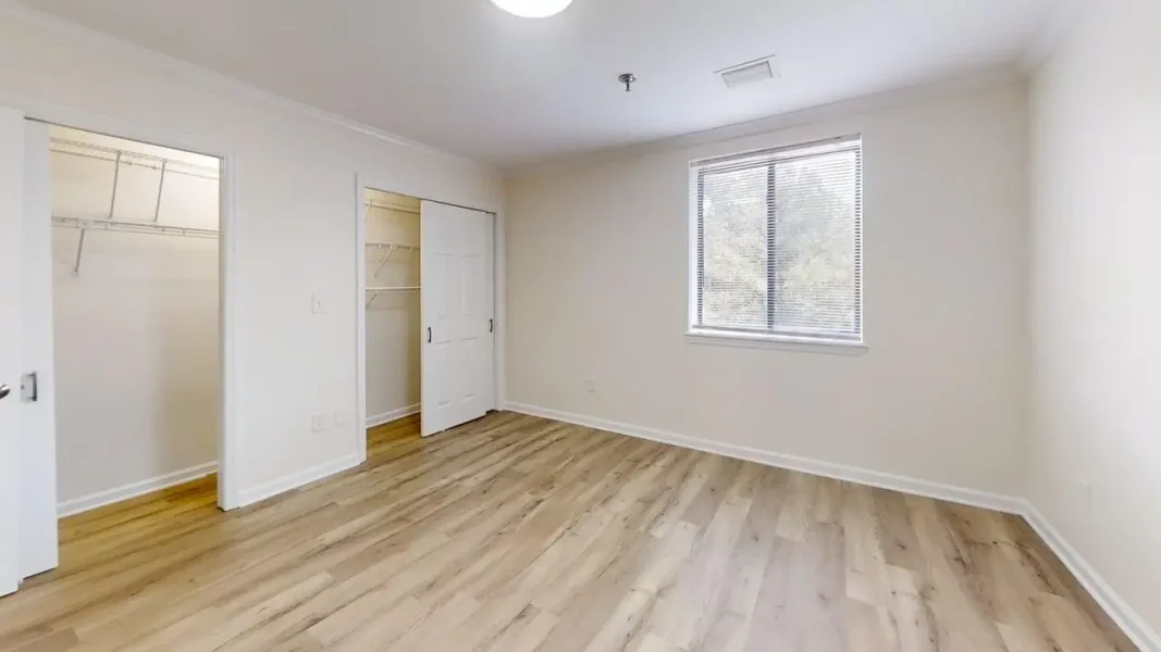 Bright empty bedroom with light wood flooring, a window with blinds, and a dual-door closet.