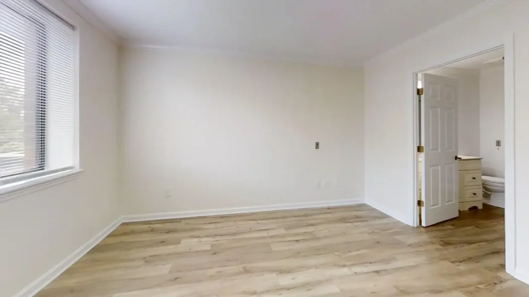 Bright empty room with light wood floors, a large window with blinds on the left, and an open doorway to a bedroom
