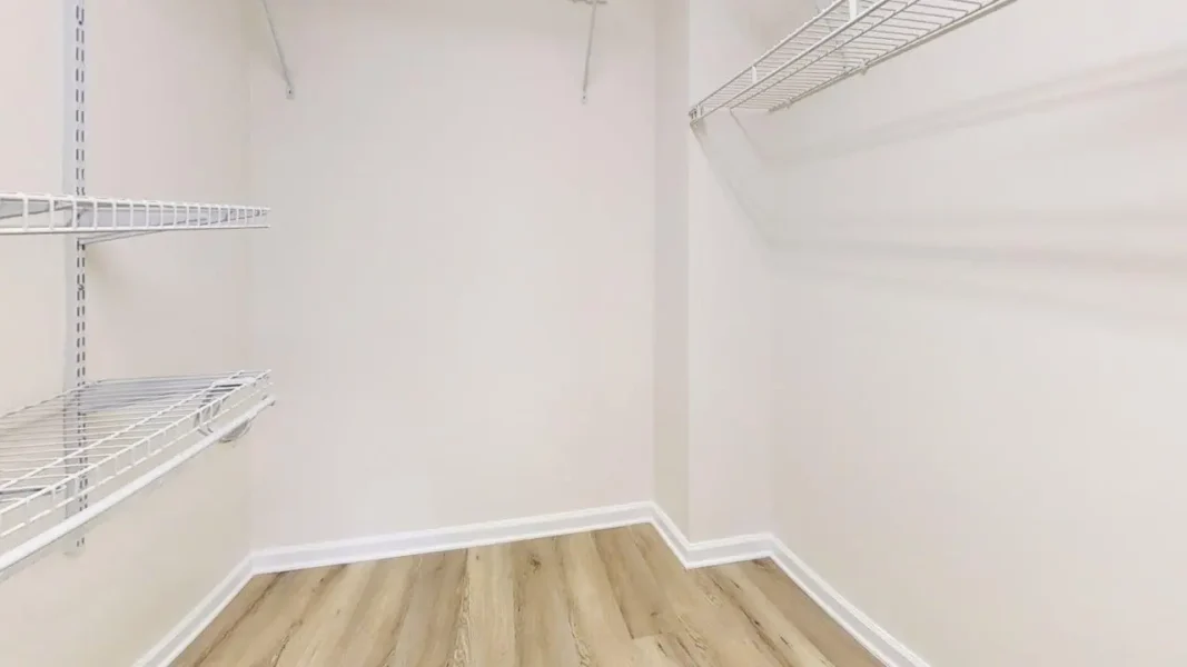 Empty walk-in closet with white wire shelves on both sides and light wood flooring.
