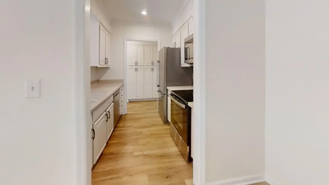 Narrow white kitchen with white cabinets, stainless steel fridge and oven, and light wood flooring