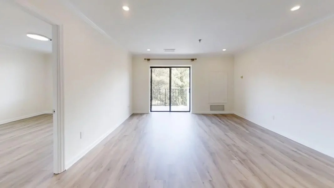 Bright empty living room with light wood floors, white walls, and a sliding glass door opening to a balcony.