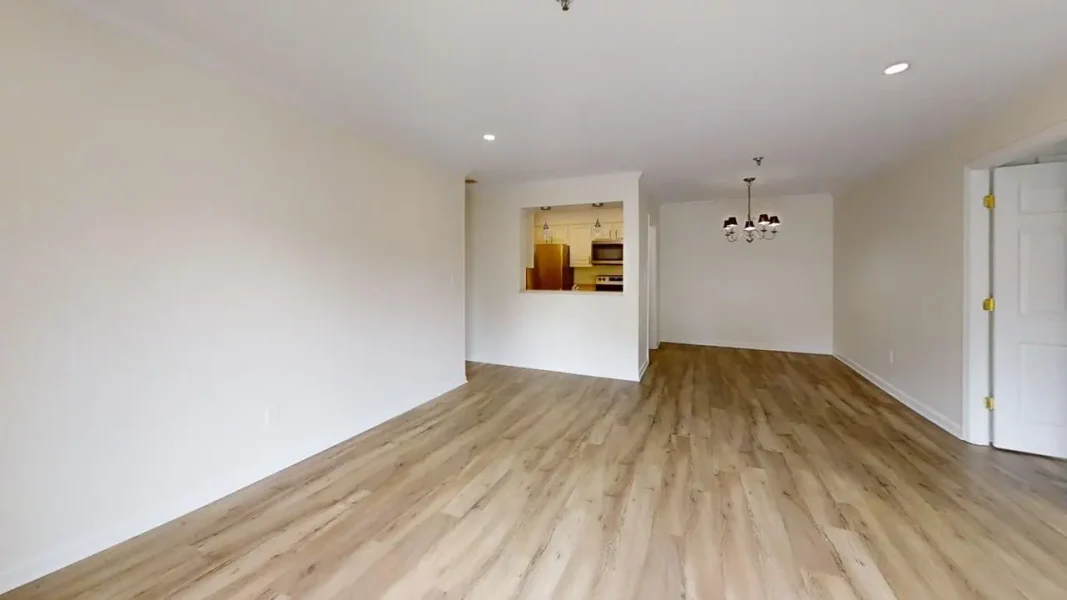 Empty open-plan living and dining area with wood-look flooring, white walls, and a pass-through window to the kitchen; a chandelier in the dining nook.