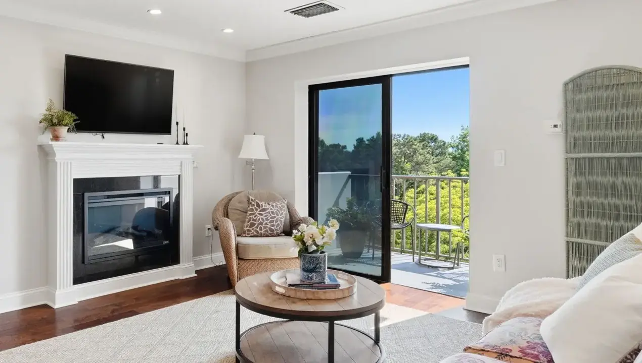 Living room with a white mantel fireplace and wall-mounted TV, a wicker chair, and a sliding glass door leading to a balcony.
