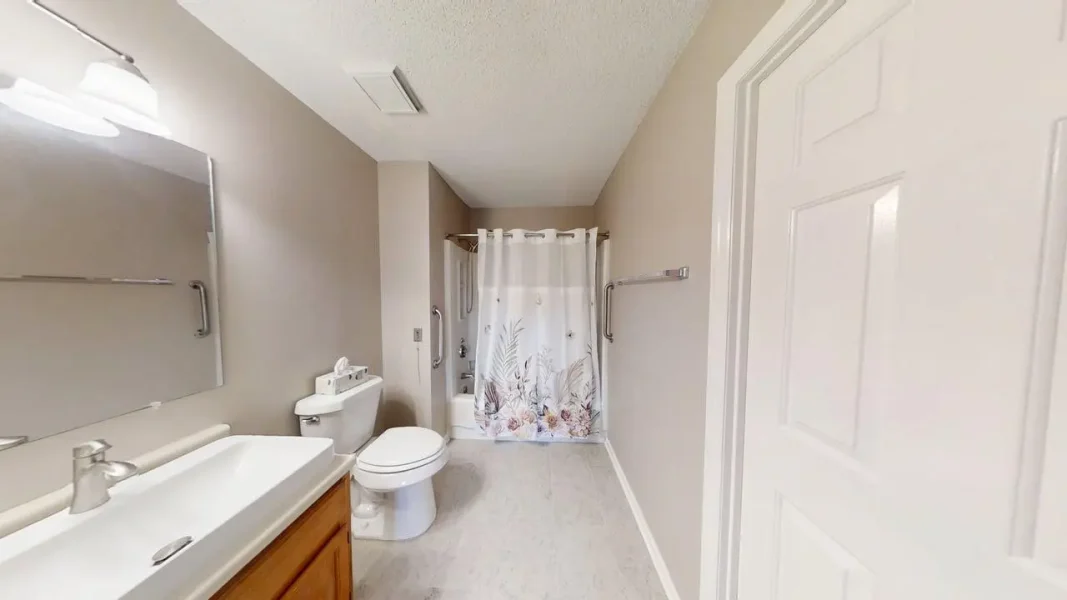 Bathroom interior with a white sink, wooden vanity, and a large mirror on beige walls, plus a toilet nearby and a floral-patterned shower curtain in the tub/shower area.