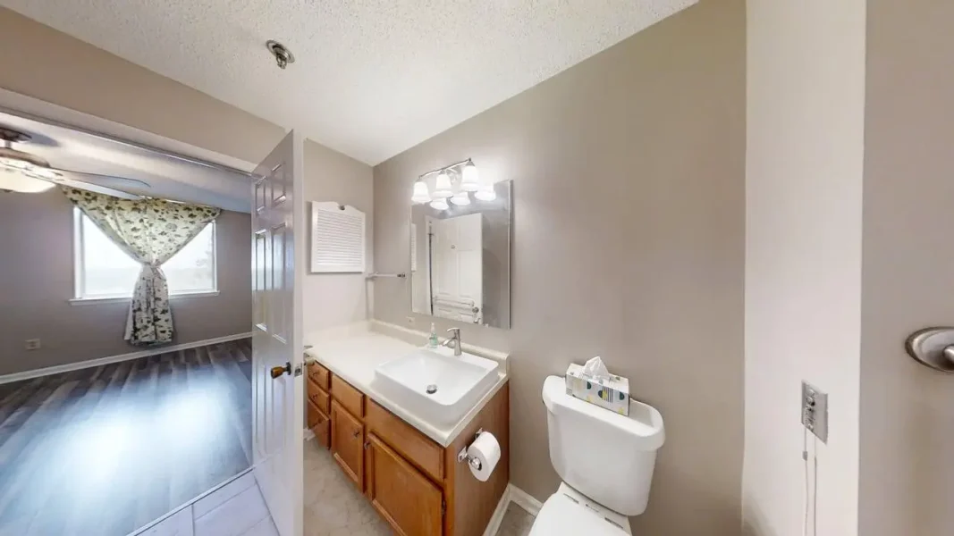 Bathroom with wooden vanity, white sink, mirrored cabinet, and toilet, open to a bedroom with floral curtains.