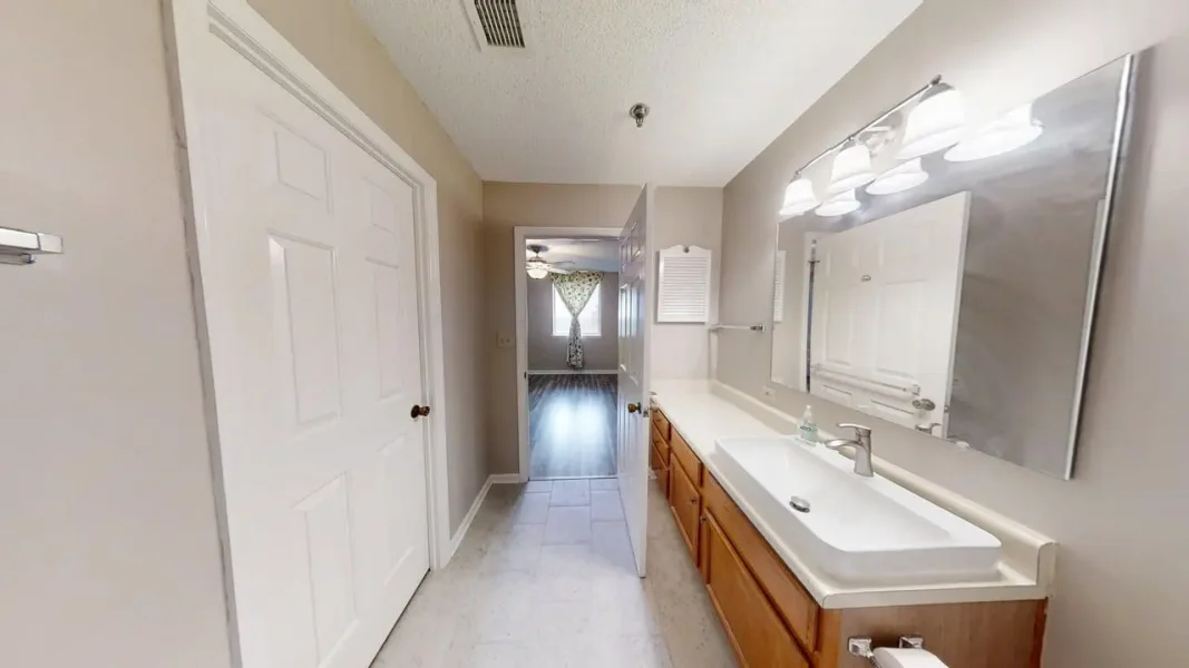 Bathroom with white rectangular sink on a wooden vanity, large wall mirror, and bright overhead lights; doorway ahead to a sunny room.