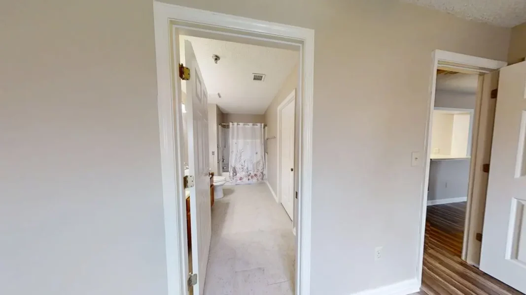 View into a bathroom with a floral shower curtain, toilet, and vanity along a light-colored tiled floor in a beige hallway.