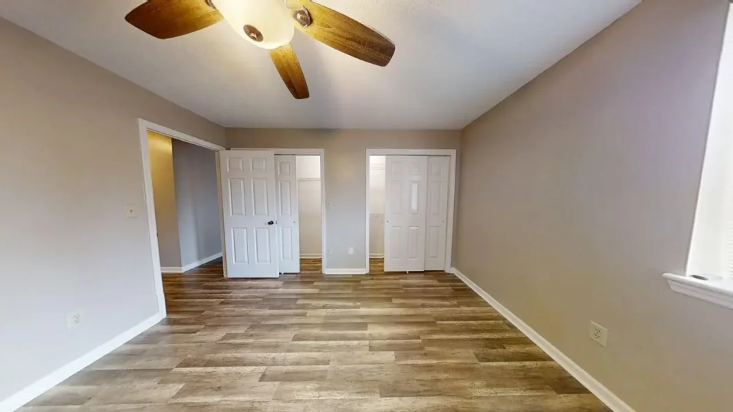 Empty bedroom with wood-look flooring, beige walls, and a ceiling fan; two white closet doors and a doorway to a hall on opposite wall; window on the right.