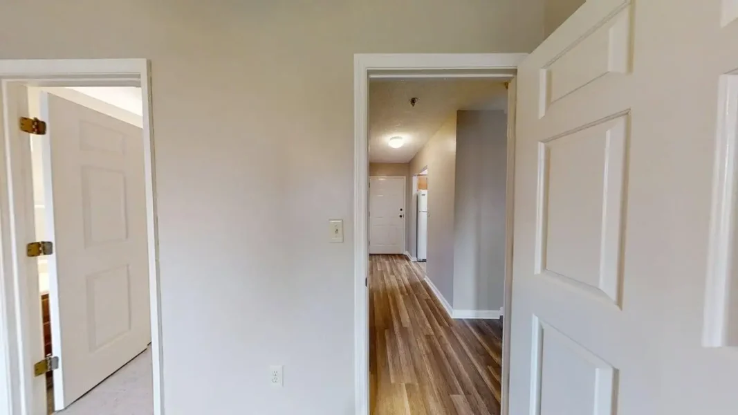 Interior view of a home: a short hallway with wood laminate flooring, light gray walls, white baseboards, and open doors on both sides.