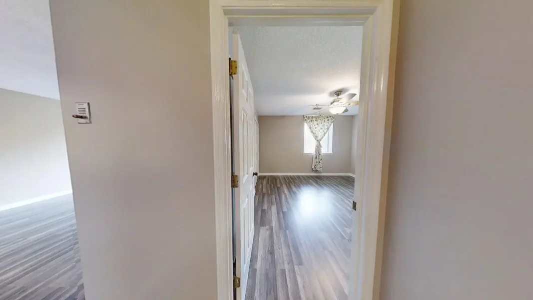 Empty living room viewed from a doorway: gray walls, wood-look flooring, a window with a floral/ patterned curtain, and a ceiling fan.