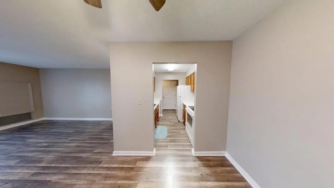 Open living area with wood-look flooring and beige walls, doorway to a small kitchen with white appliances visible in the back. - Cooking area seen through a central doorway from a spacious living room. - Spacious living room flowing into a compact galley-style kitchen with white fridge and cabinets.