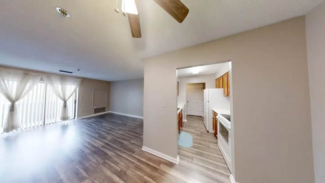 Bright living room with laminate flooring, sheer curtains, and an opening to a small kitchen.
